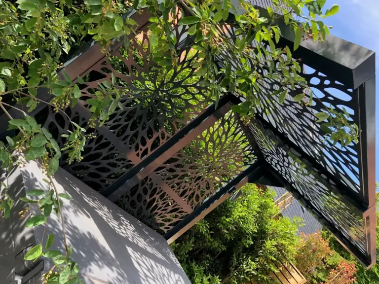 Close-up view of a custom-designed decorative metal roof panel with intricate floral patterns, providing shade and elegance in a garden setting.